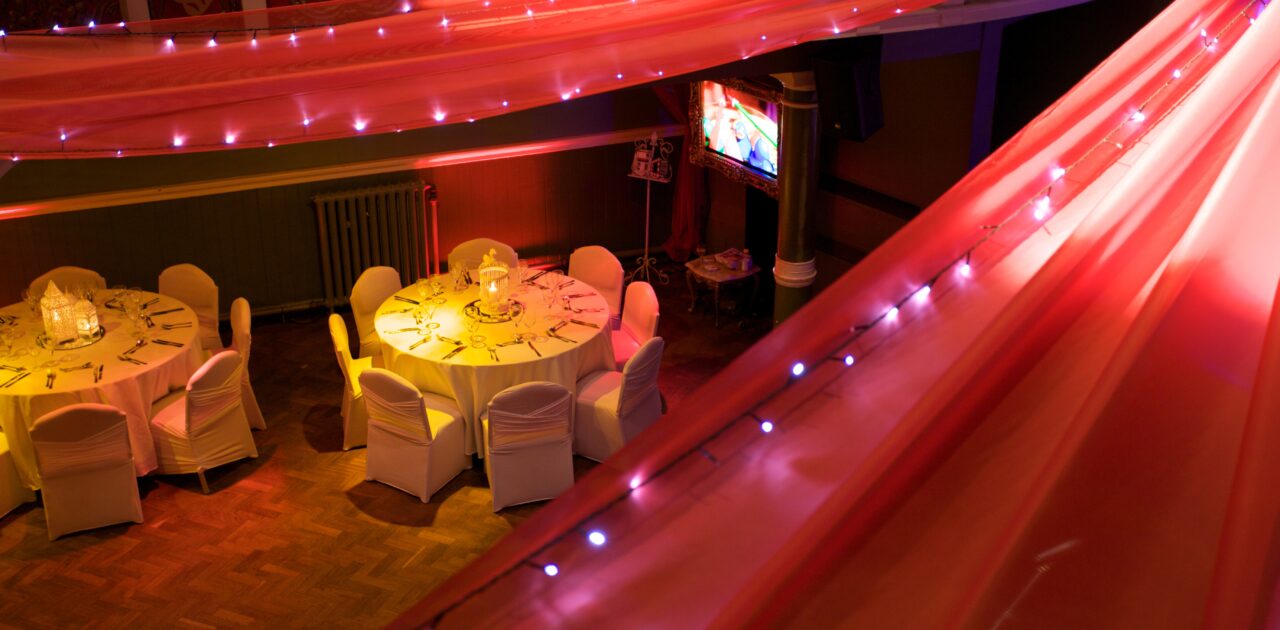 Red fabric and twinkling lights hang from a central point in the ceiling and drape down to the balcony. Between the fabric you can see two sets of round tables and chairs, covered in white and set for silver service.