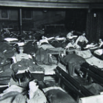 A black and white photograph of the theatre filled with young men sleeping or sitting up in rows of camp beds, the old theatre bench seating pushed to one side.