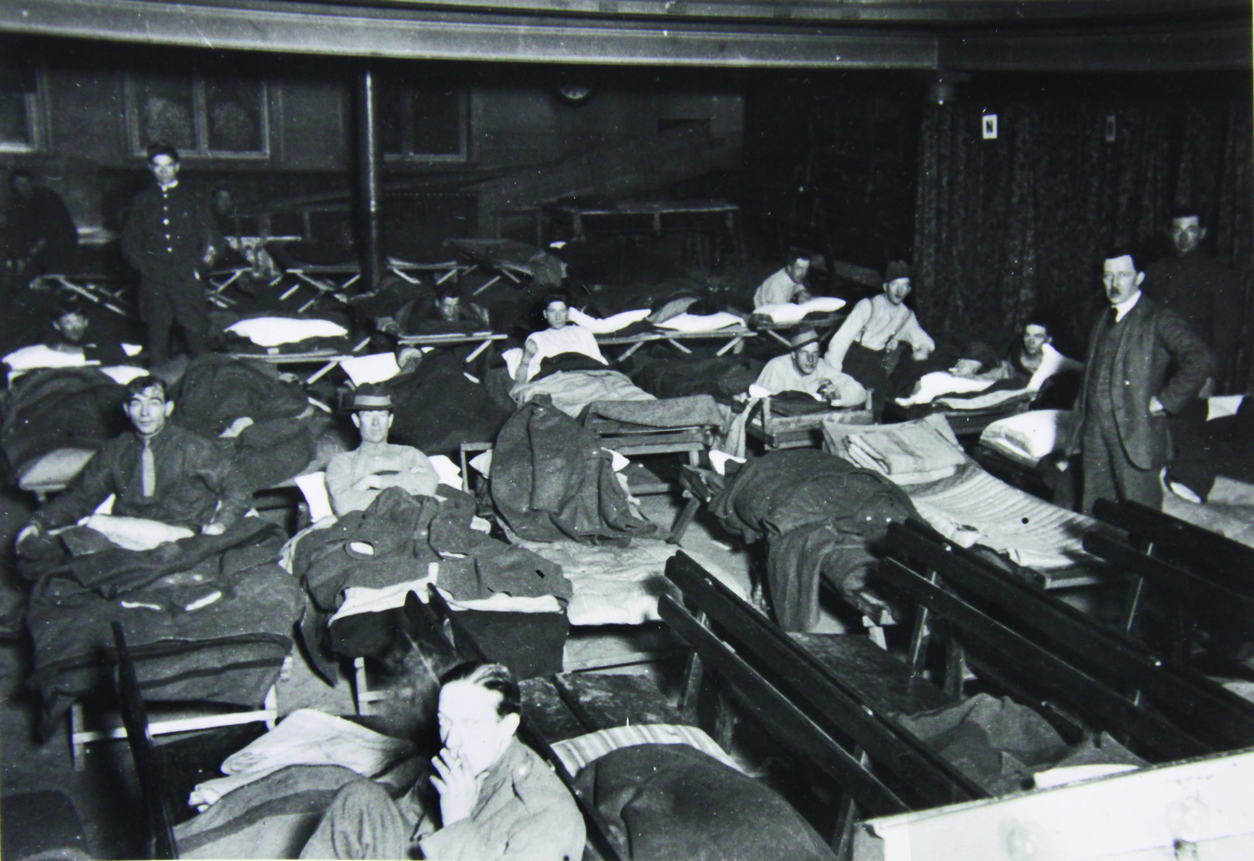 A black and white photograph of the theatre filled with young men sleeping or sitting up in rows of camp beds, the old theatre bench seating pushed to one side.
