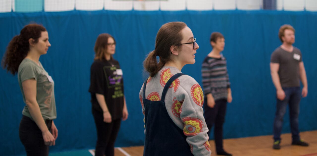 A divider is pulled across the room with a basket ball net visible beyond it. A group of people stand in a circle, looking to someone off camera