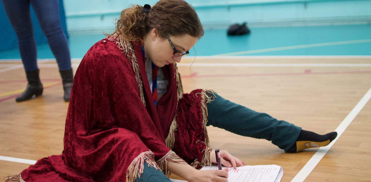 A woman sits on the floor of the sports hall, writing in a script