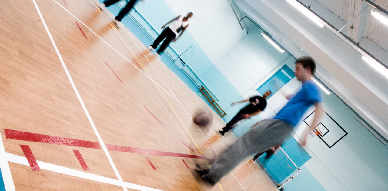 A slanted photo of a group playing football in the sports hall. In the backgroup we see a basket ball net as well as floor markings for various sports.