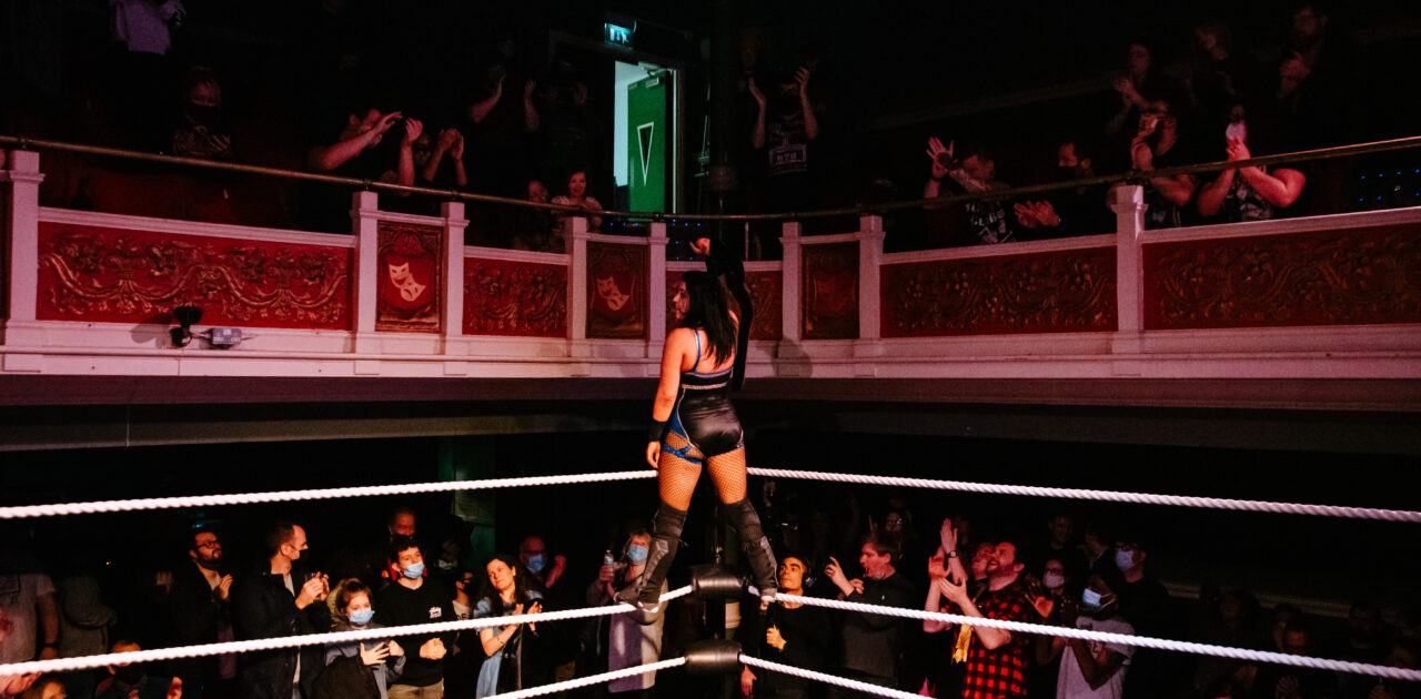 A wrestling ring sits on the floor, inside the ede of the balcony. A female wrestler stands on the ropes, holding the winners belt high to a cheering crowd.