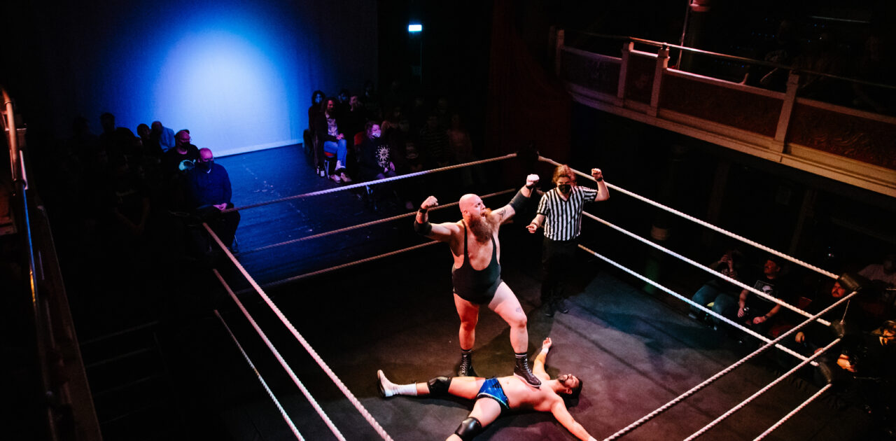A wrestling ring sits on the floor, inside the ede of the balcony., the stage behind it. The audience sit on the stage with a walkway down the middle, and around the ring. A male wrestler is splayed on the floor of the ring, another with a foot on his chest, arms aloft in celebration, the referre gesturing towards him as the winner.