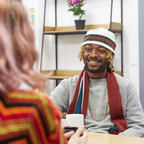 A young man and young woman sit opposite each other with mugs of tea, chatting.