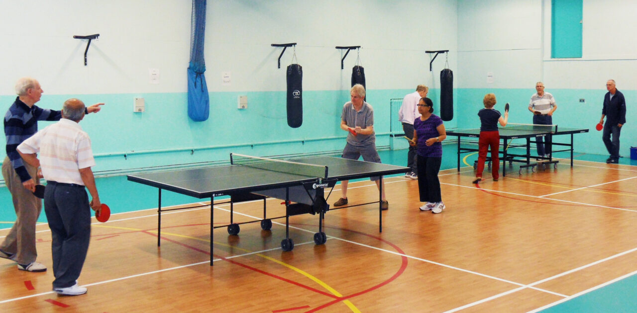 A mixed group of older people play across two table tennis tables. We can see punching bags along the back wall and the dividing net packed away against the wall.