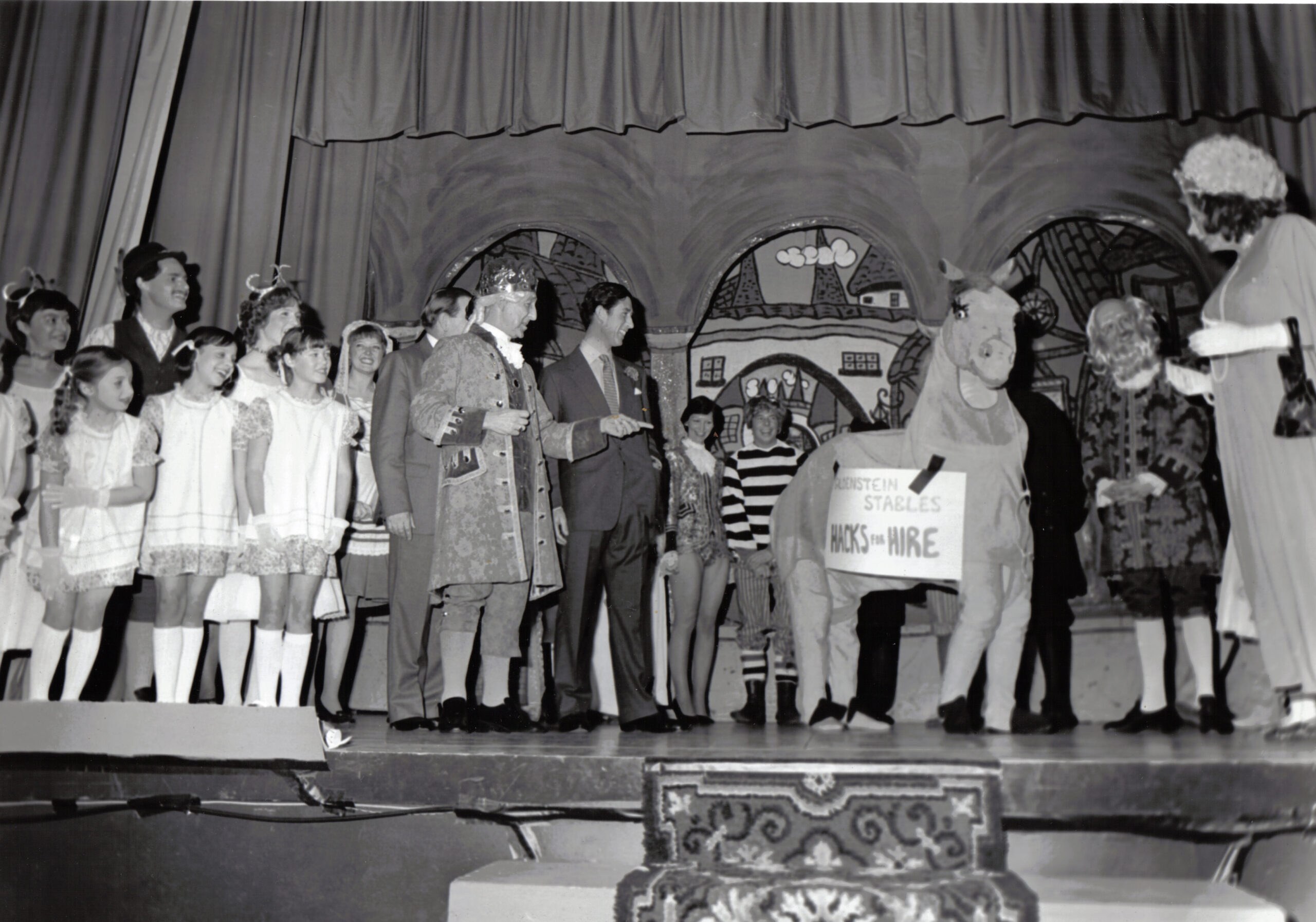 A black and white photo of HM King Charles III on the Theatre stage with the panto cast, including the youth chorus, king and a pantomime horse.